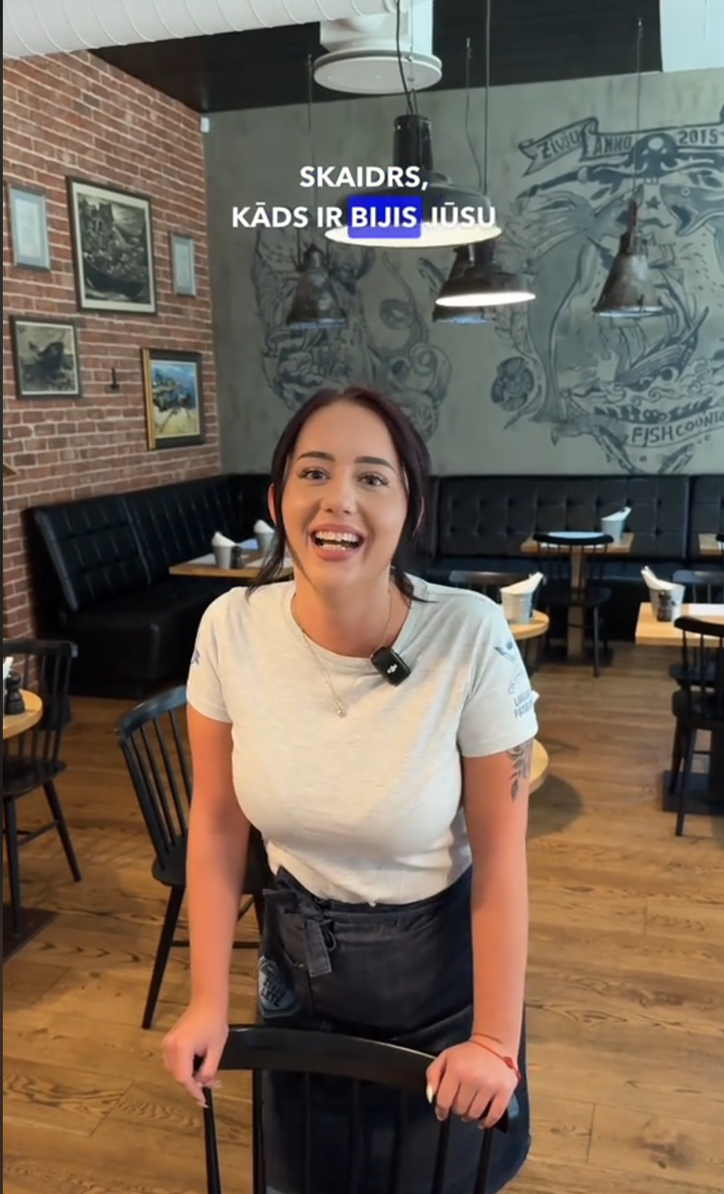 Simpātiska jauna sieviete baltā kreklā un tumšā priekšautā restorānā